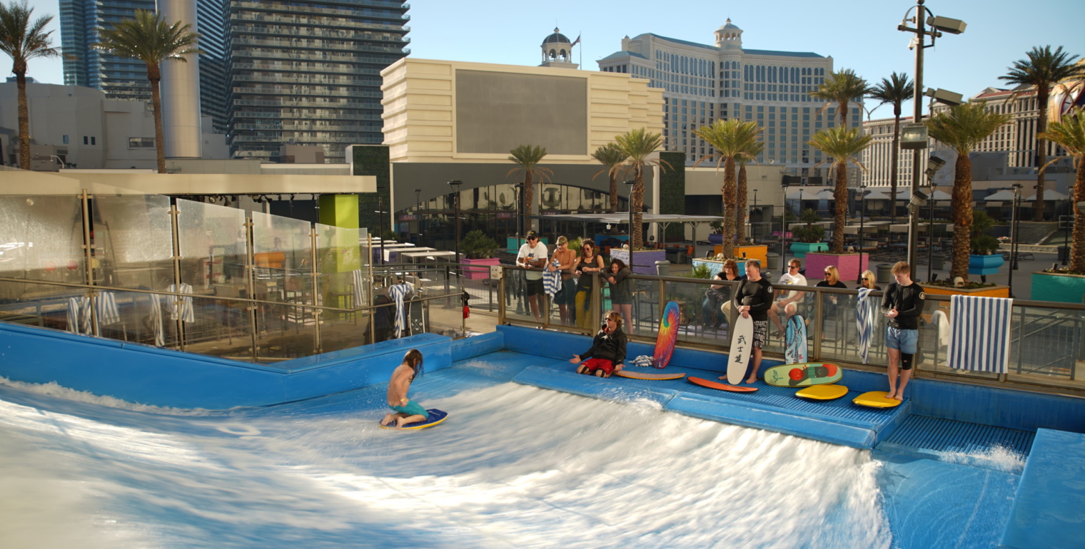A group of people watch a teenager kneeling on a surfboard trying to surf